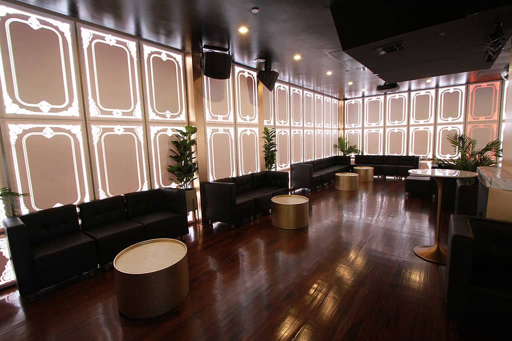 The image depicts an interior view of a modern, spacious lounge or bar area with black leather seating, a polished wood floor, and a large window with white blinds.